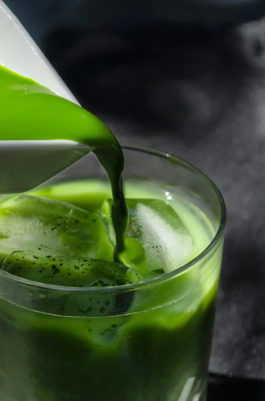 A thick stream of whisked matcha poured into a glass of ice, the surface still foaming.