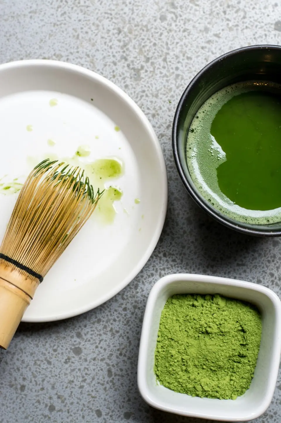 Seen from above: a bamboo chasen resting on a white plate, a bowl of bright green whisked matcha, and a small dish of dry powder.
