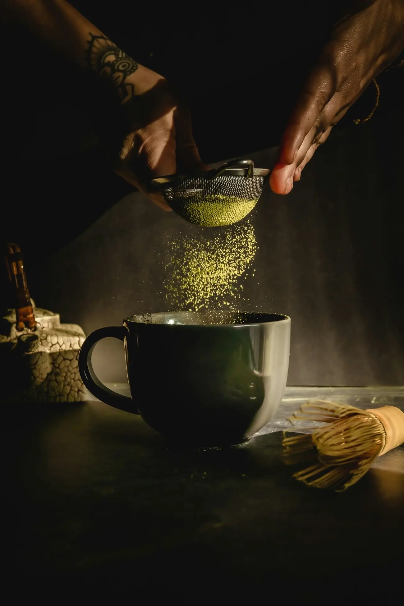 Matcha being pushed through a mesh strainer into a dark mug, the falling powder lit from behind.