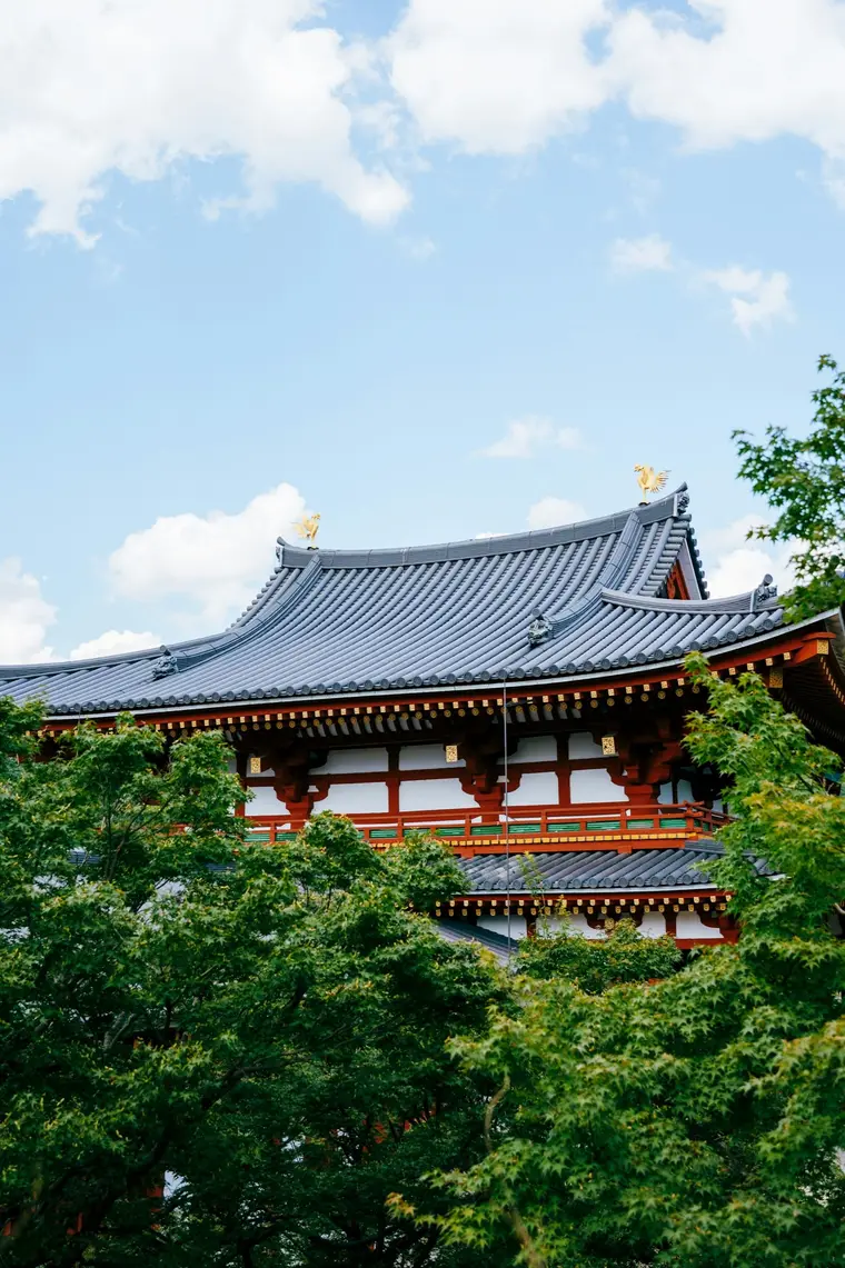 A vermilion and white Japanese temple hall with a sweeping tiled roof, framed by green trees under a blue sky.