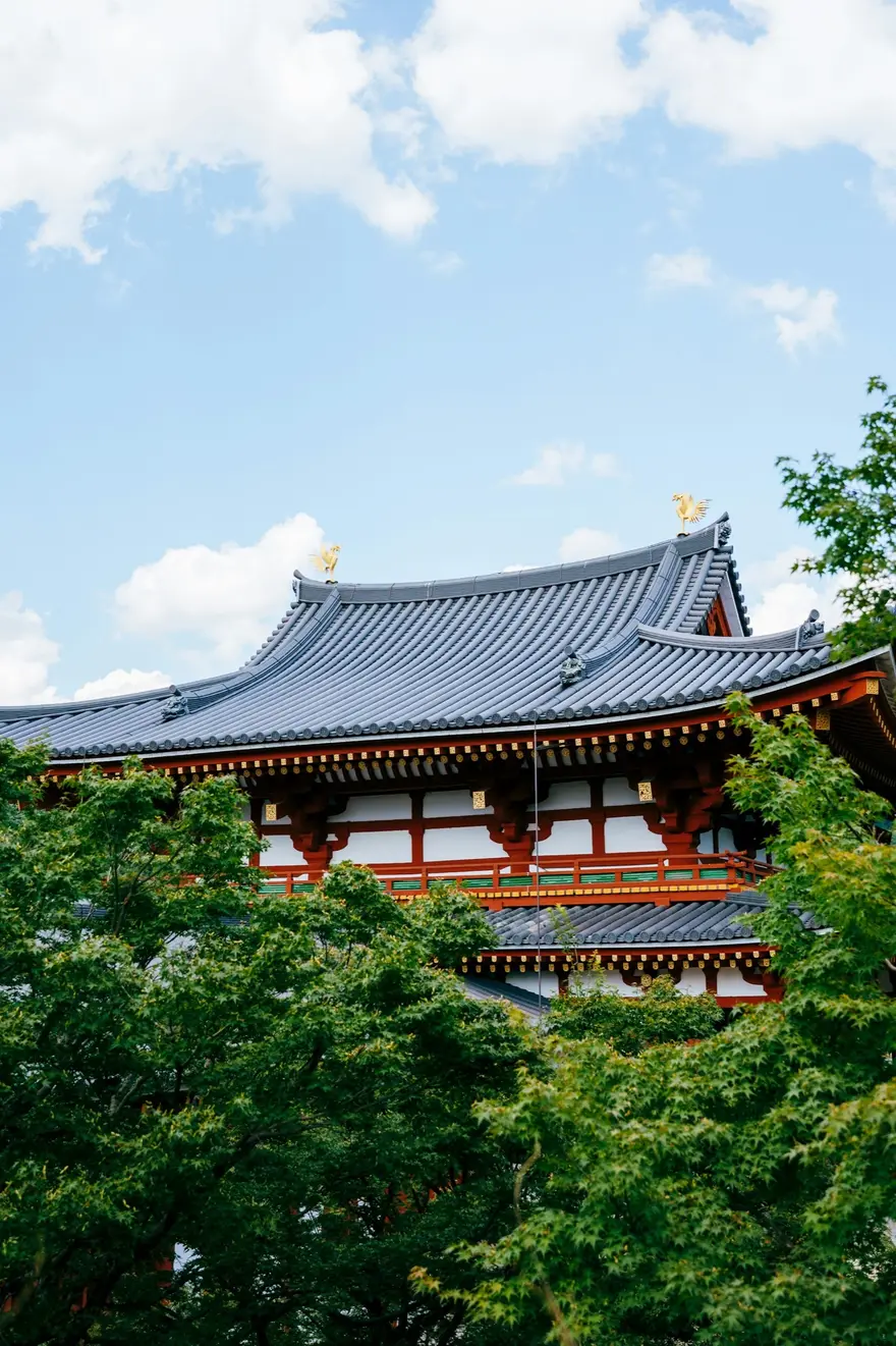 A vermilion and white Japanese temple hall with a sweeping tiled roof, framed by green trees under a blue sky.