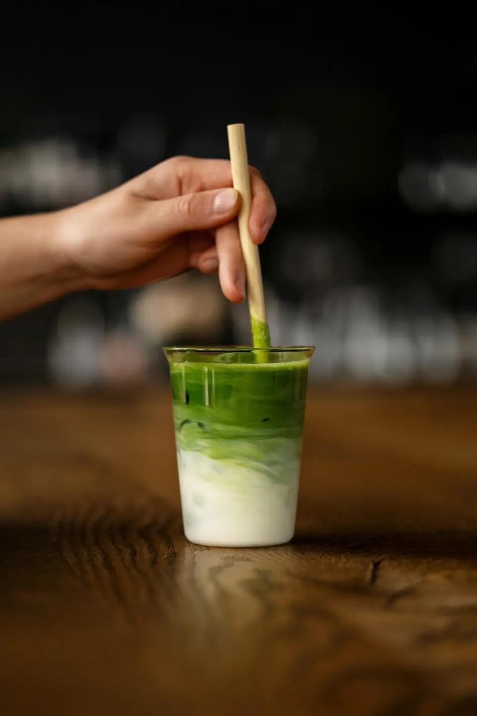 A hand stirring a layered iced matcha, the green drawing down through the milk.